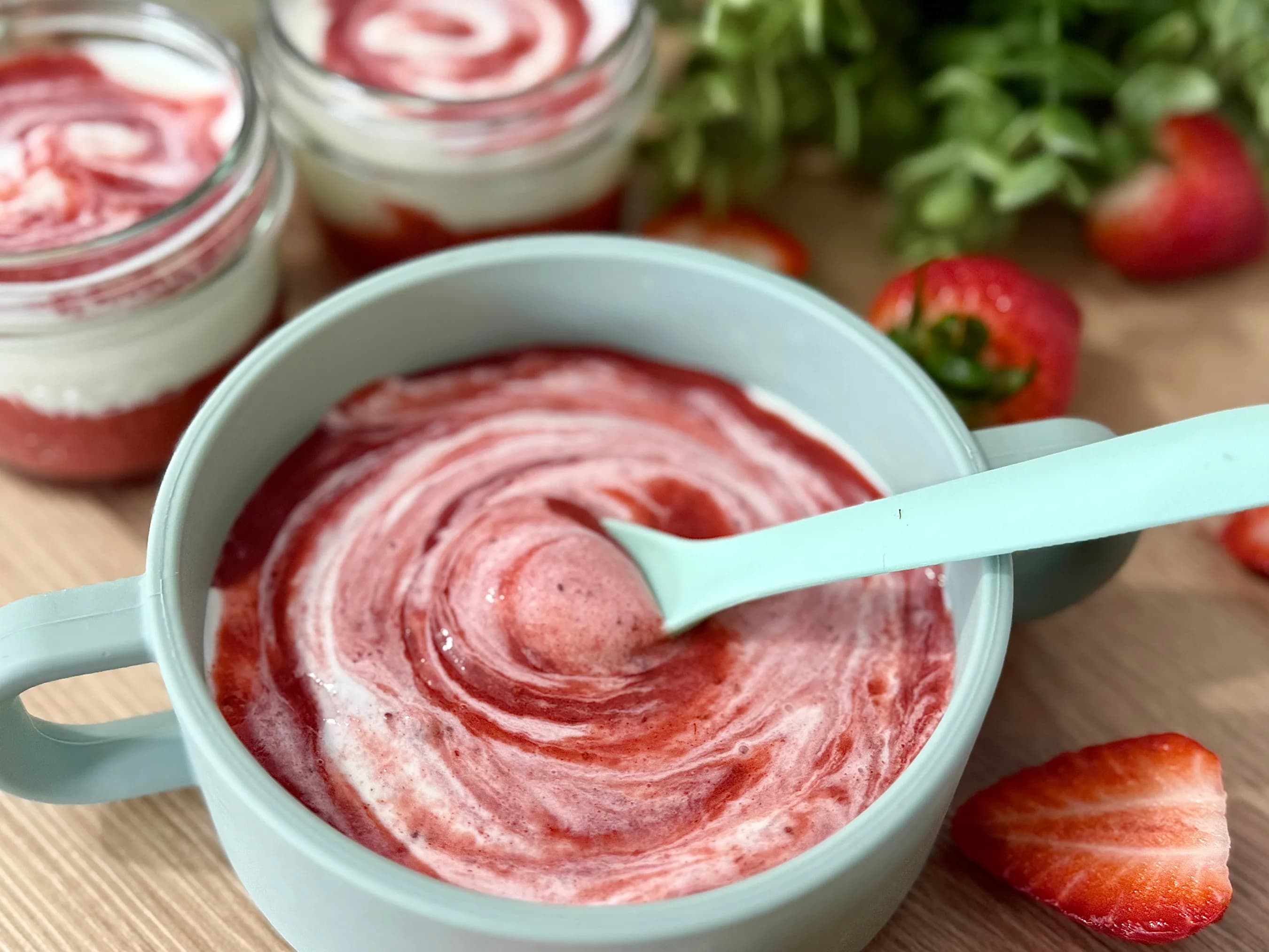 "Close-up of a blue bowl filled with creamy strawberry yogurt swirled with red fruit puree, with a mint green spoon. In the background, two glass jars of layered strawberry yogurt parfaits and fresh strawberries are visible. Ideal for breakfast, a healthy snack, or baby food."