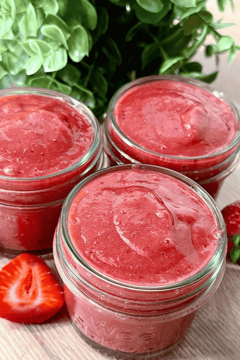 Three small glass jars filled with a smooth, vibrant homemade pink strawberry jam, surrounded by fresh strawberries on a light wooden surface.