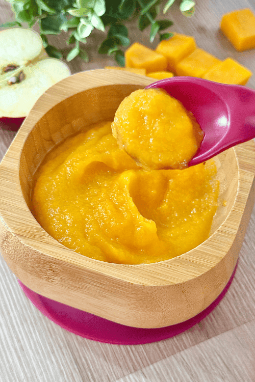 Smooth squash and apple puree served in a wooden bowl with a baby spoon, surrounded by fresh squash cubes and apple slices.
