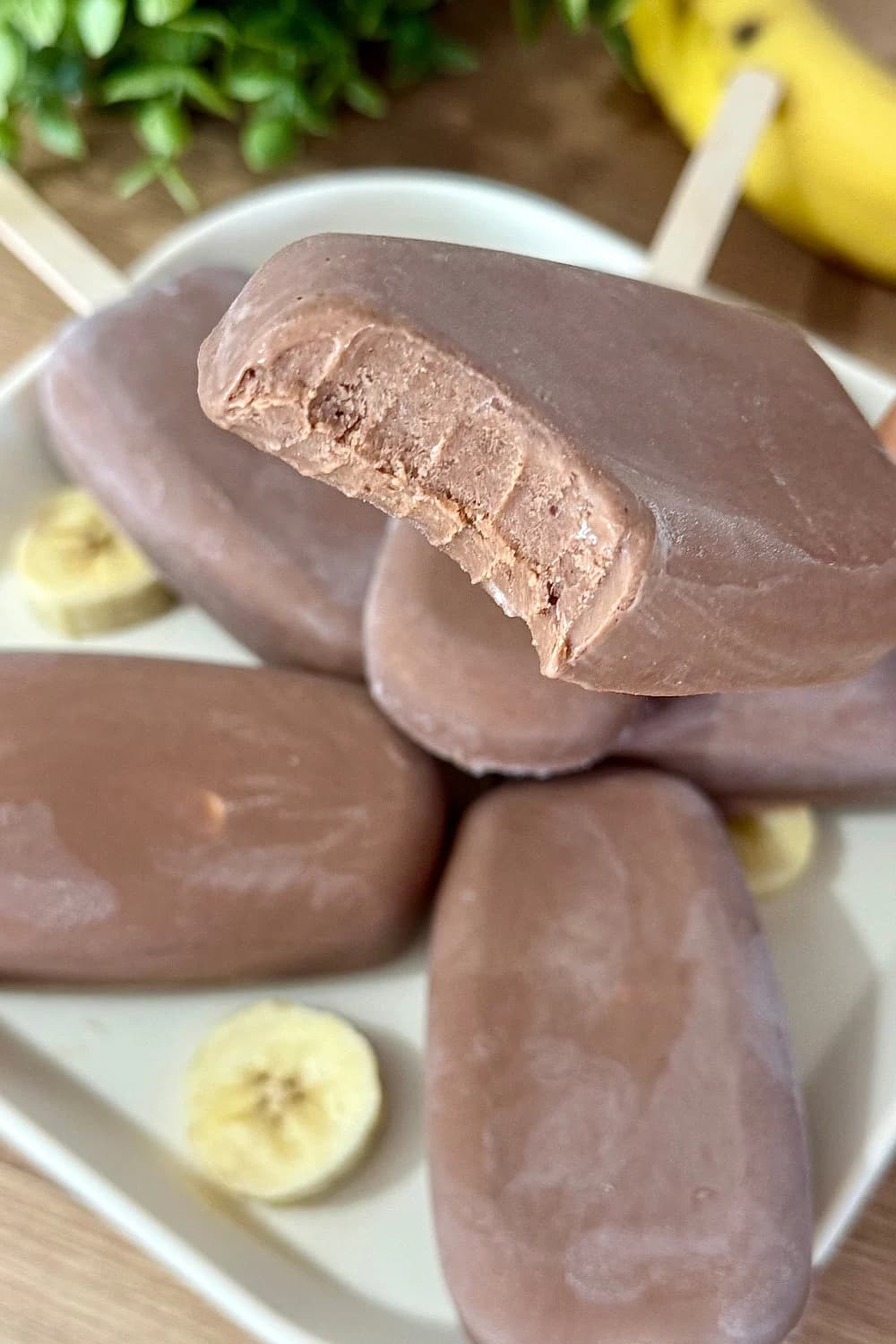 Close-up of a creamy chocolate popsicle with a bite taken out, showing its smooth texture. Other chocolate popsicles and banana slices are blurred in the background on a white plate. Ideal for a refreshing frozen dessert.