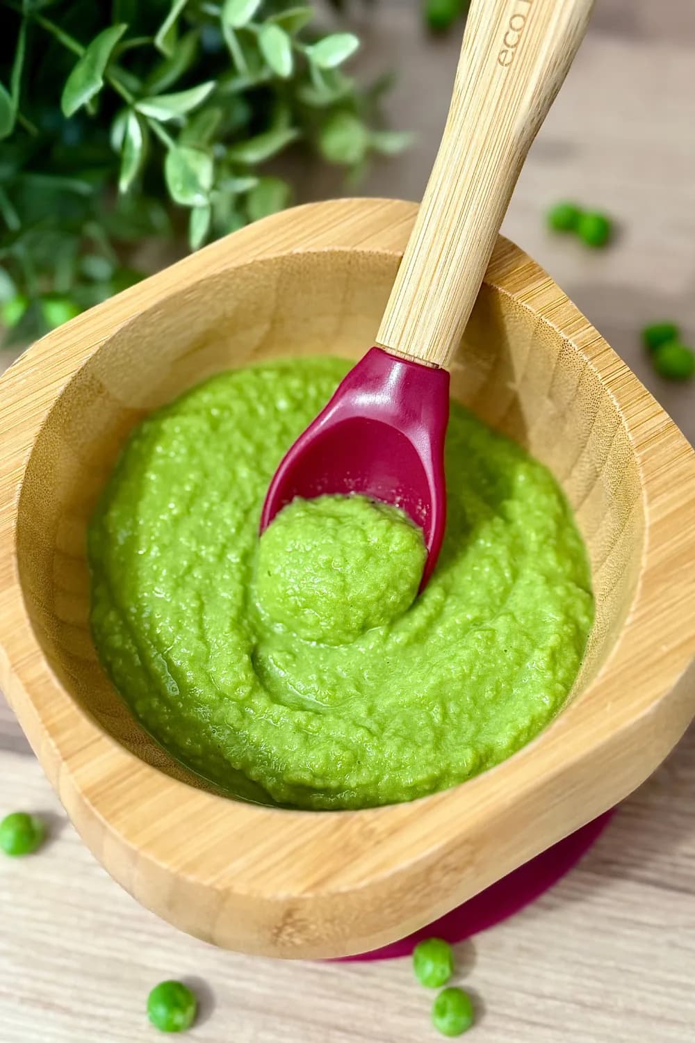 Close-up of smooth, vibrant green homemade pea puree in a natural bamboo baby bowl, with an Eco Rascals spoon scooping a portion. Loose peas and green foliage are in the background, illustrating a healthy and easy-to-prepare first food for babies 6 months and up.