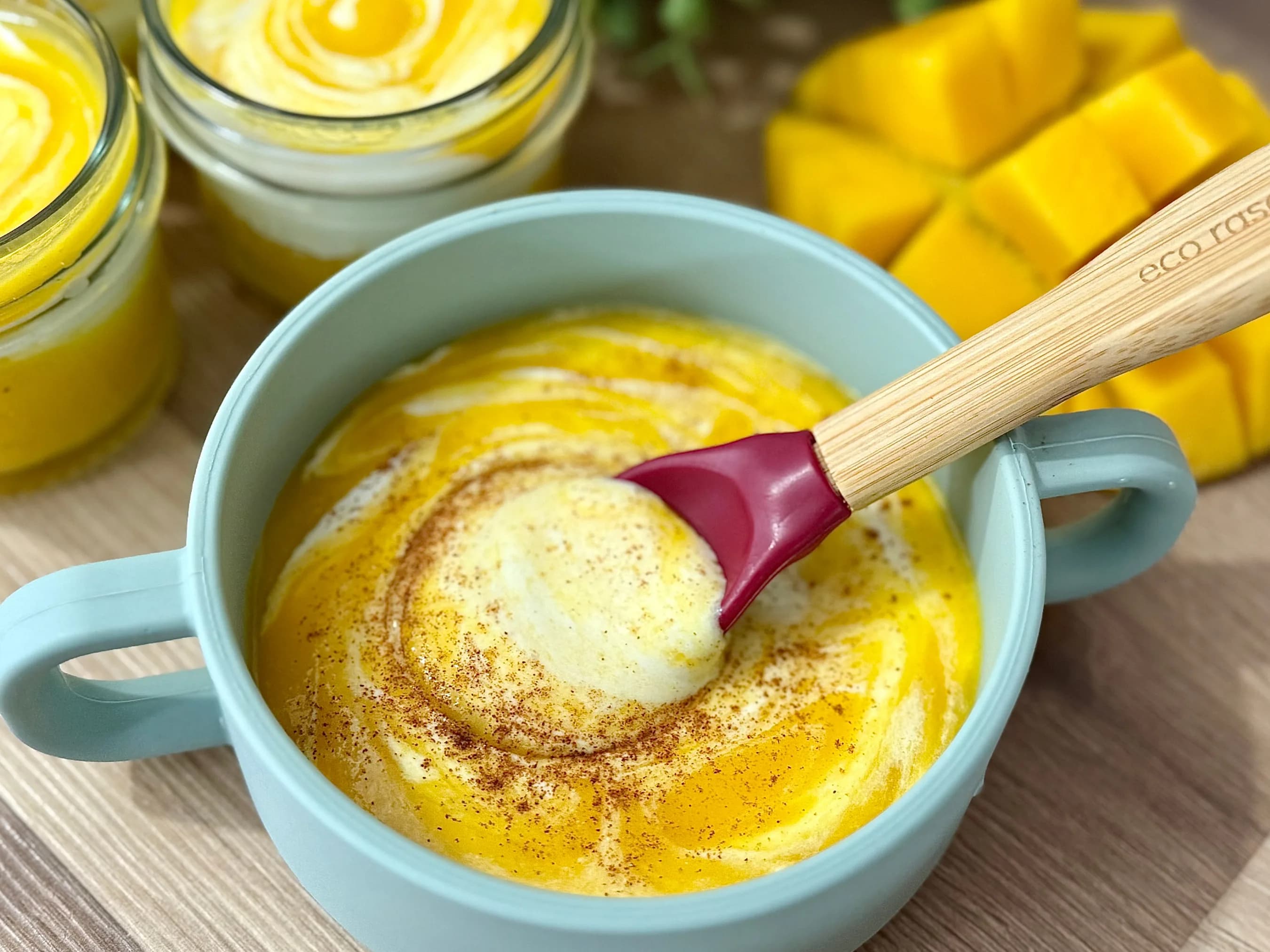 Homemade mango yogurt in a child-friendly blue bowl with an Eco Rascals bamboo spoon, swirled with cinnamon, indicating a delicious and healthy dessert or baby-led weaning food. Individual layered mango yogurt jars and fresh diced mango are in the background.