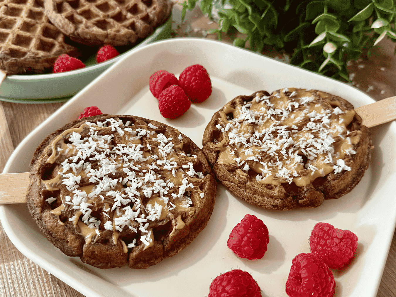 Coconut chocolate waffle pops topped with nut butter and shredded coconut, served on a plate with fresh raspberries.