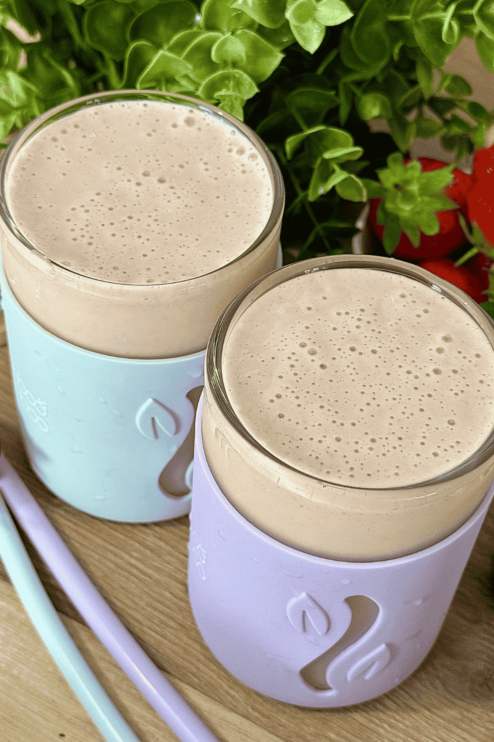 Two glasses of creamy strawberry banana smoothie in pastel-colored sleeves, surrounded by fresh strawberries and green leaves on a wooden surface.
