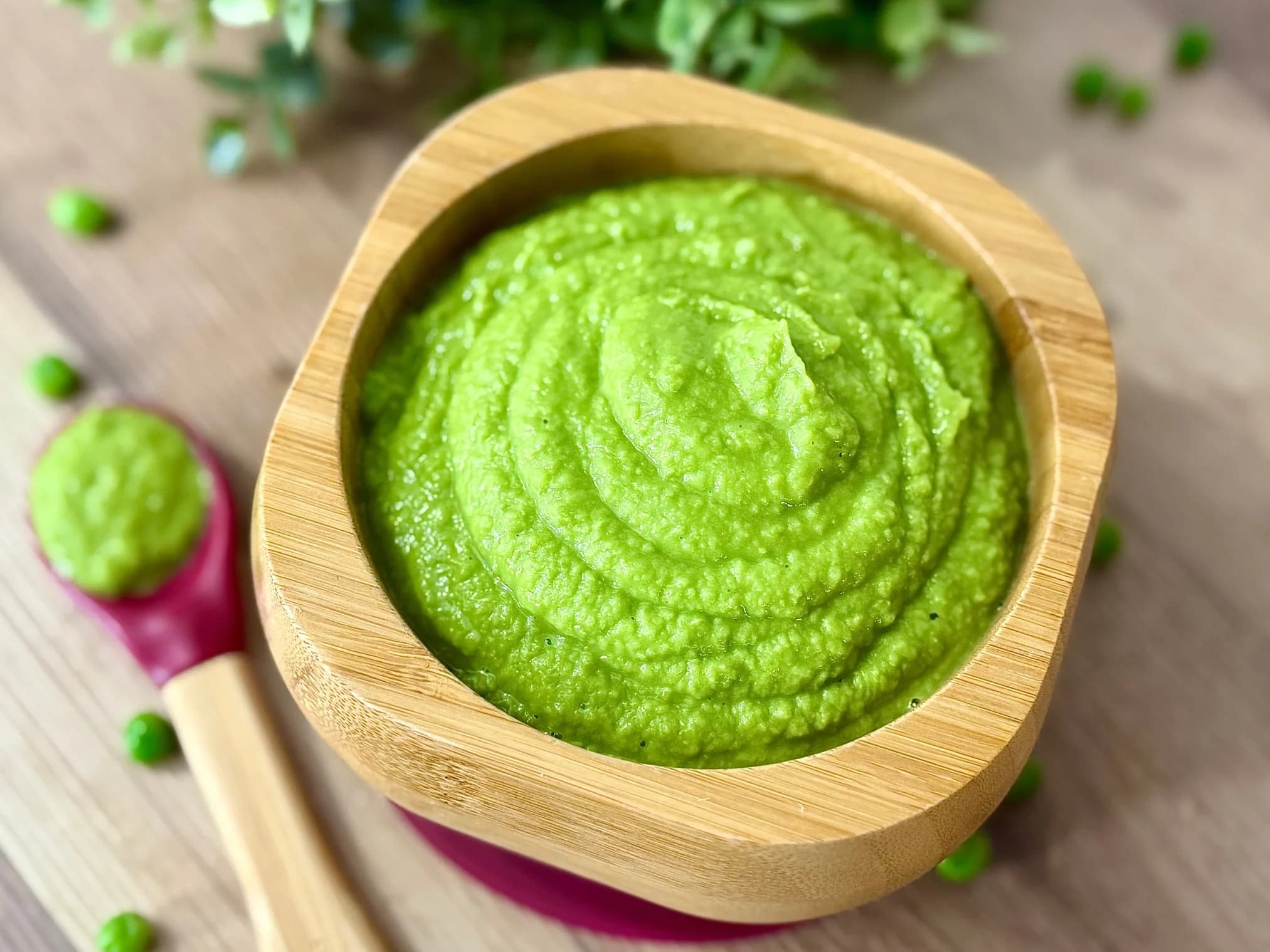 Vibrant green, smooth homemade pea puree in a natural wooden baby bowl with a bamboo spoon, ready for feeding. Scattered fresh peas and green foliage in the background highlight this healthy, easy-to-digest first food for infants and toddlers.