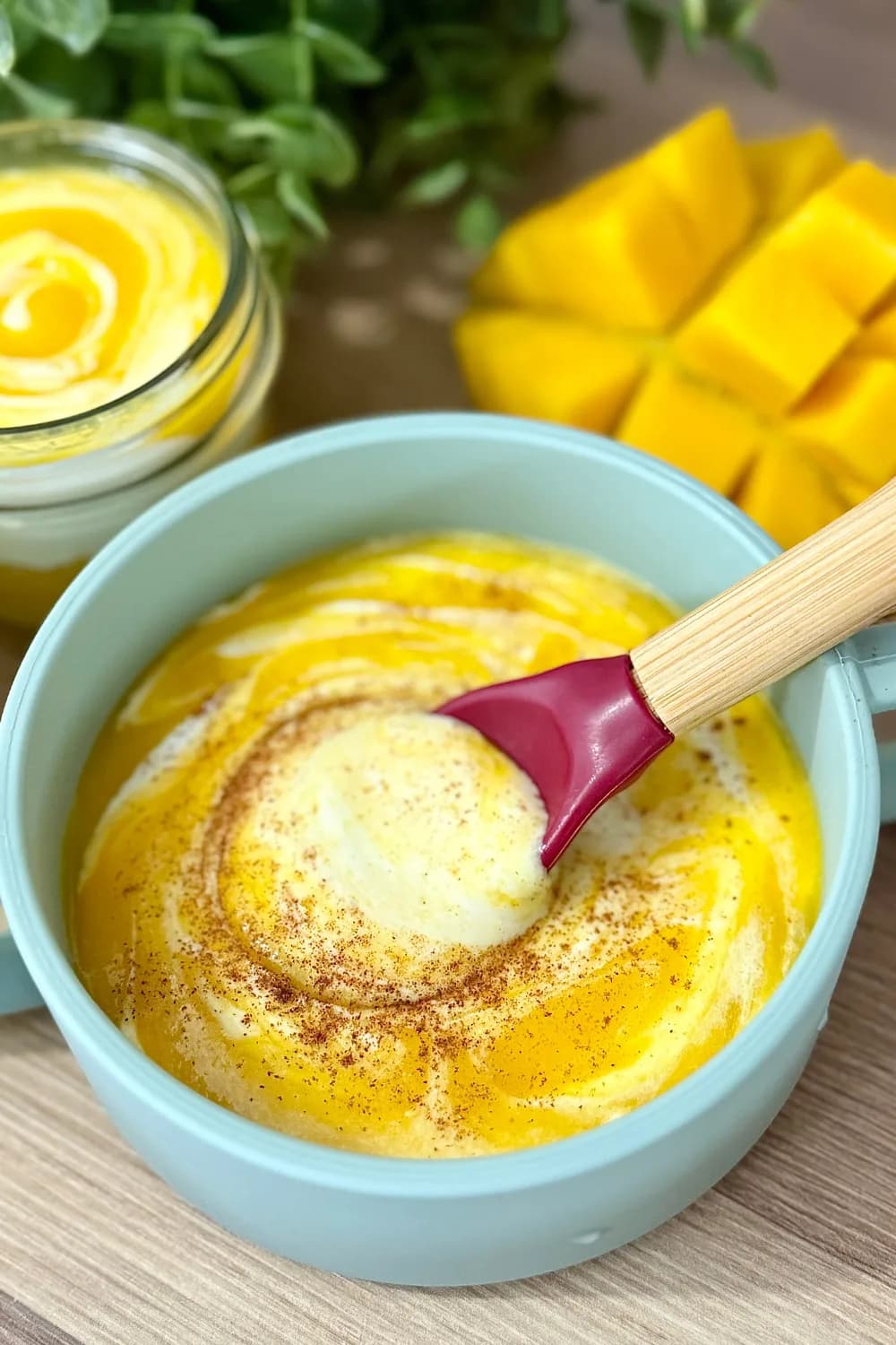 Close-up of creamy homemade mango yogurt in a blue baby bowl with a bamboo spoon, swirled with a sprinkle of cinnamon. In the background, fresh diced mango and a glass jar of layered mango and yogurt are visible. Ideal for baby's first foods or a healthy toddler snack.