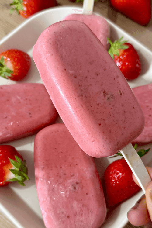 Homemade strawberry yogurt popsicles arranged on a white plate with fresh whole strawberries for garnish.