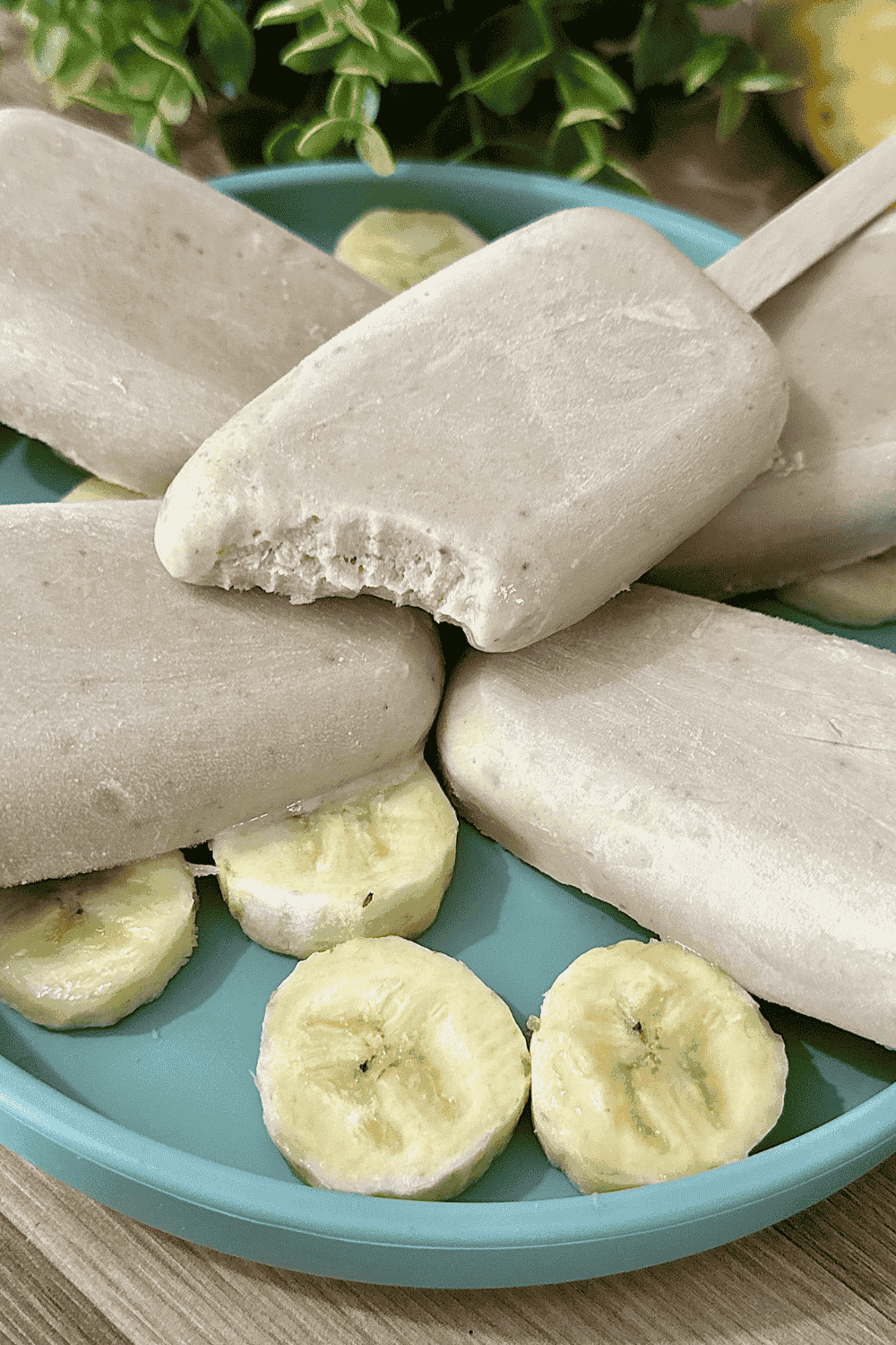 Four creamy banana popsicles arranged in a teal dish with banana slices underneath, one popsicle showing a bite taken out to reveal its smooth texture. A fresh banana is partially visible in the background.